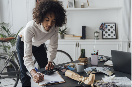 A woman leans down to write on a notepad on a coffee table