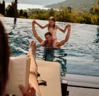 A man neck-deep in a swimming pool with his young daughter perched on his shoulders