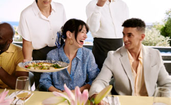 Two guests sit side-by-side at a table as a server places a dish in front of them