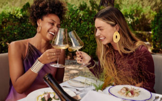 Two guests seated at a patio table share a toast with their wine glasses