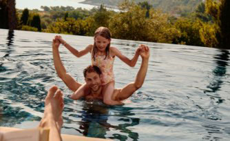 A man neck-deep in a swimming pool with his young daughter perched on his shoulders