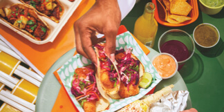 Close-up of a hand reaching for a taco and another hand scooping a tortilla chip into a side of salsa