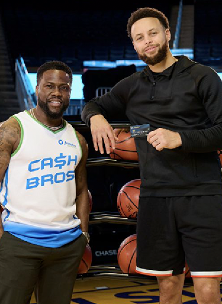 Stephen Curry stands courtside holding a Freedom Unlimited Credit Card next to Kevin Hart