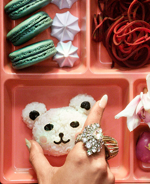 A hand places a bear-shaped Japanese rice ball into a bento box filled with sweets