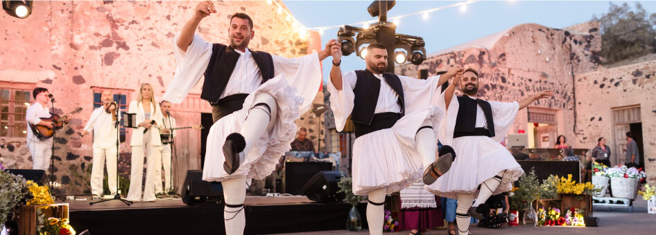 Dancers in customary clothing at an outdoor festival with a rustic stone building in the background.