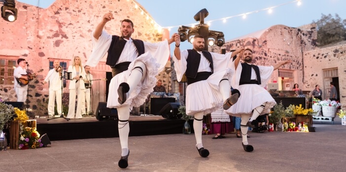 Dancers in customary clothing at an outdoor festival with a rustic stone building in the background.