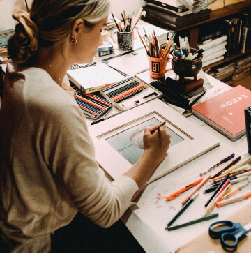 An overhead shot of an artist sitting at her desk, working on a piece with colored pencils