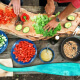 An overhead shot of Native American hands preparing peppers, avocados, greens, and dressing on an outdoor setting