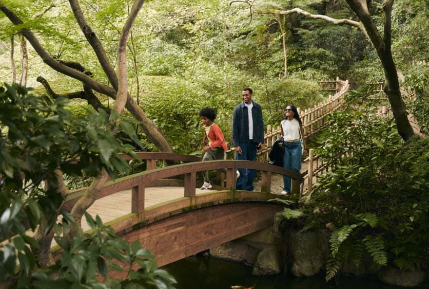 Three people walking on a wooden bridge in a lush, green forest or garden in Japan.