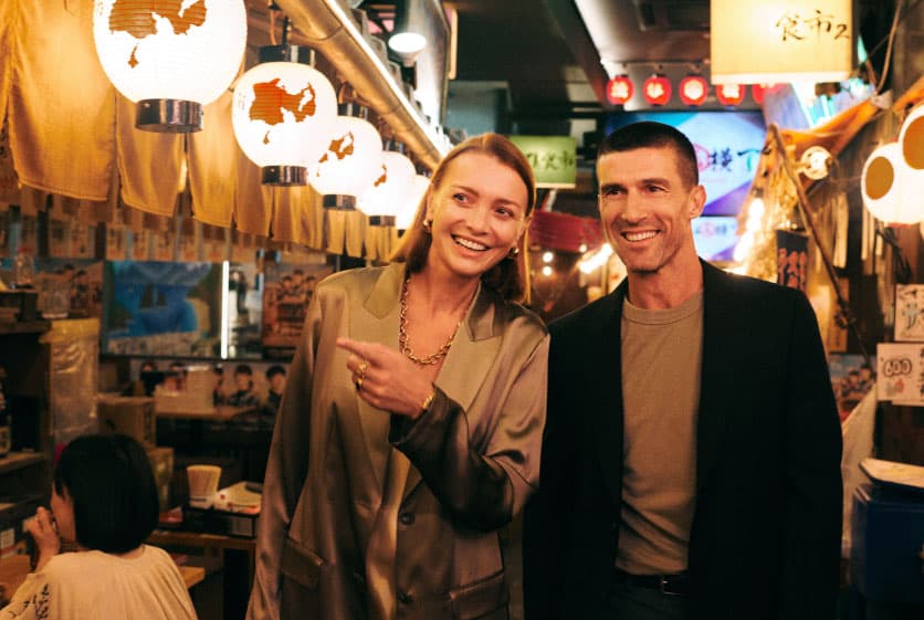 Two people walking on a street in Japan with luggage, in front of a black luxury car.