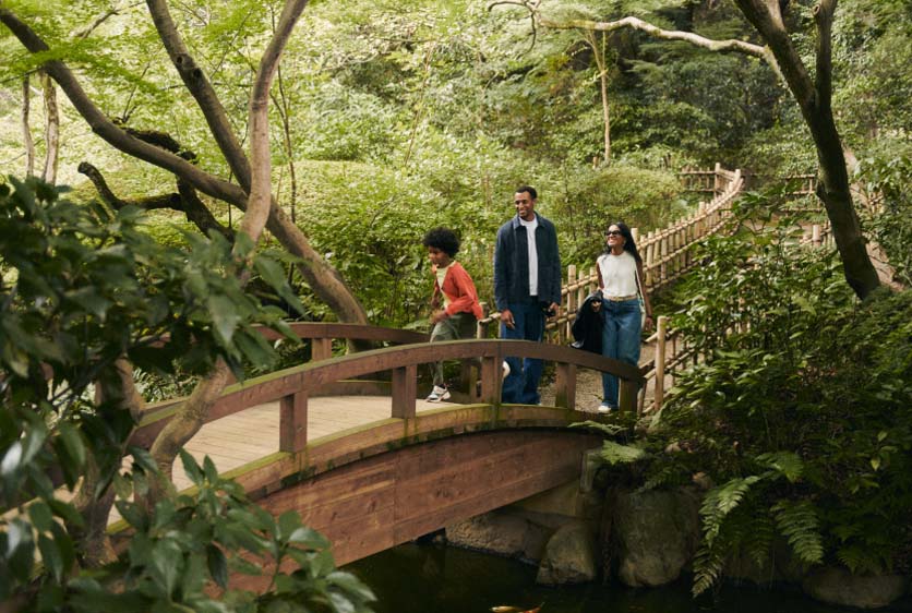Three people walking on a wooden bridge in a lush, green forest or garden in Japan.