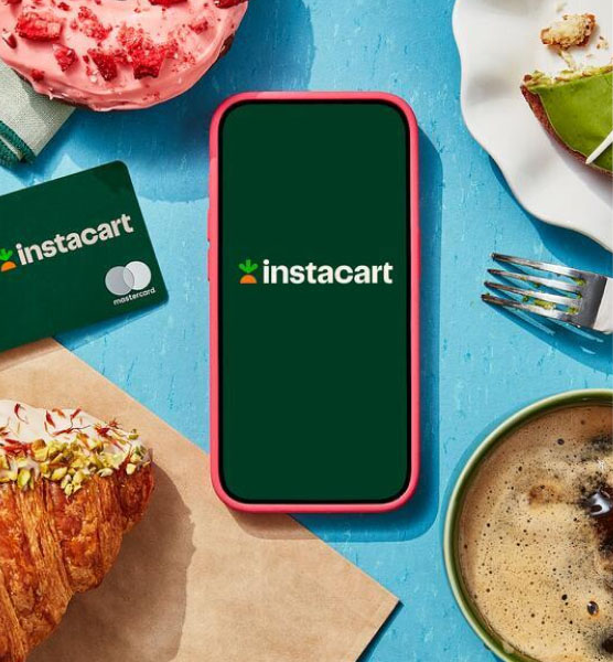 Overhead shot of smartphone with Instacart logo onscreen, surrounded by the Instacart Mastercard, colorful pastries, and a coffee.