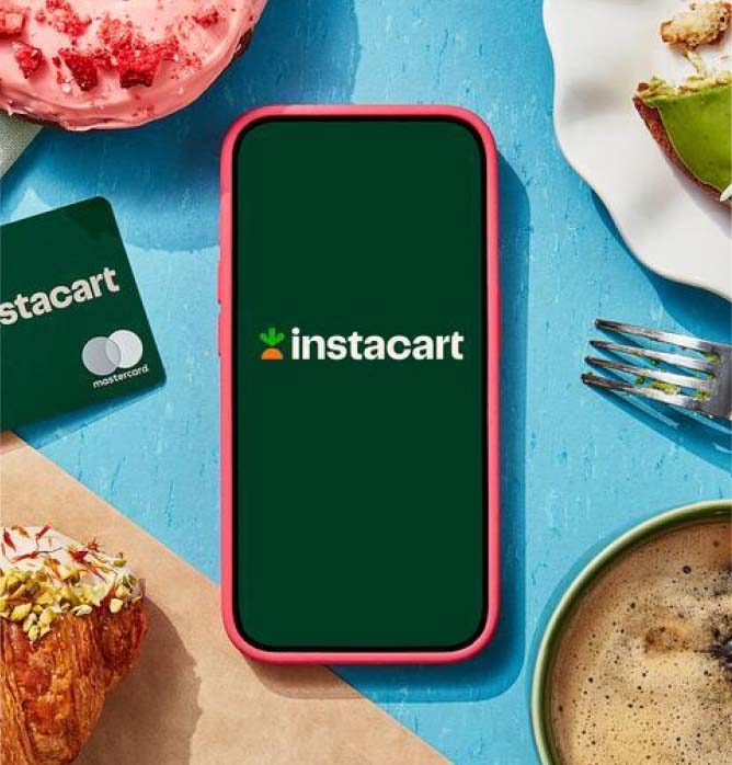 Overhead shot of smartphone with Instacart logo onscreen, surrounded by the Instacart Mastercard, colorful pastries, and a coffee.