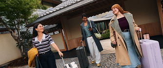 Three people walking outside a building with traditional japanese architecture, carrying luggage.