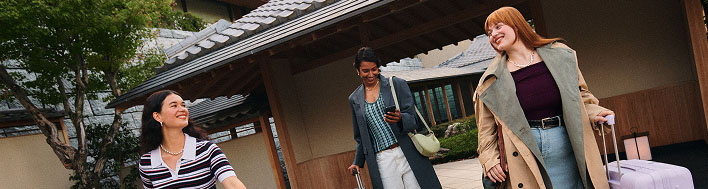 Three people walking outside a building with traditional japanese architecture, carrying luggage.