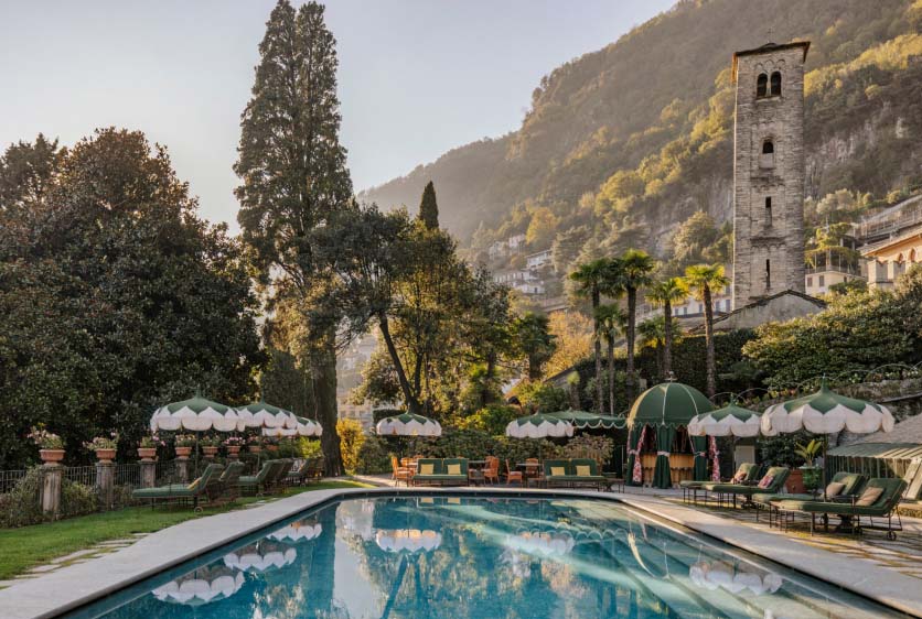 Poolside view with umbrellas, trees, and a stone tower against Lake Como hills.