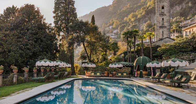 Poolside view with umbrellas, trees, and a stone tower against Lake Como hills.