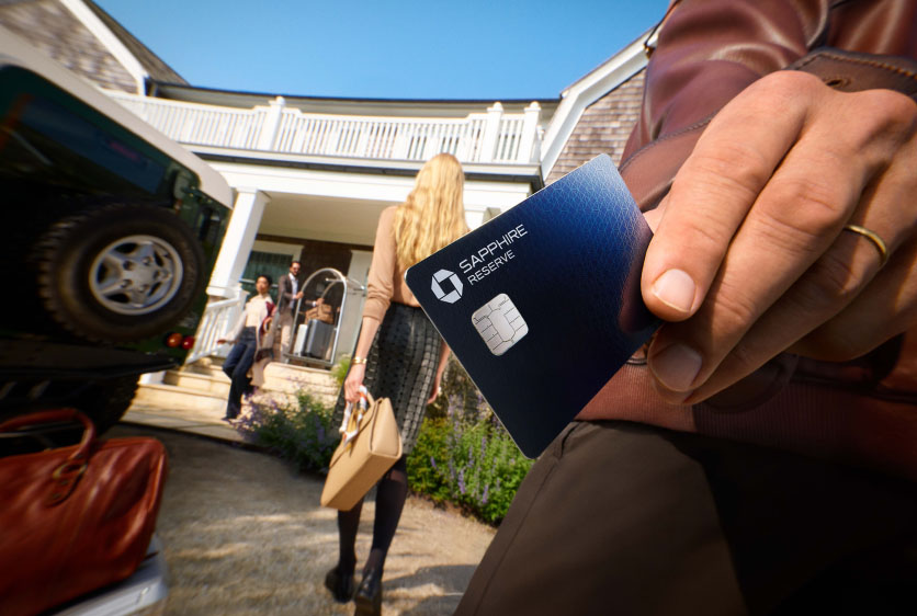 Hand holding a Chase Sapphire Reserve card with people walking toward a house.