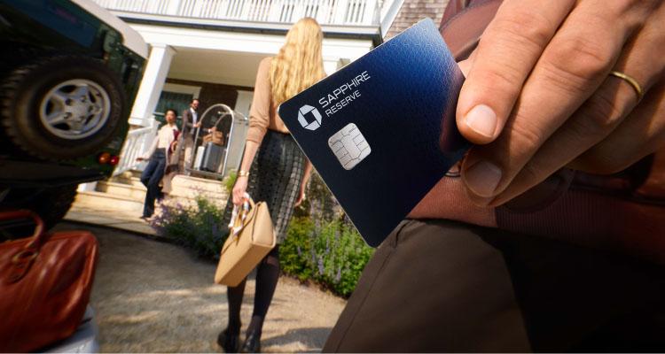 Hand holding a Chase Sapphire Reserve card with people walking toward a house.