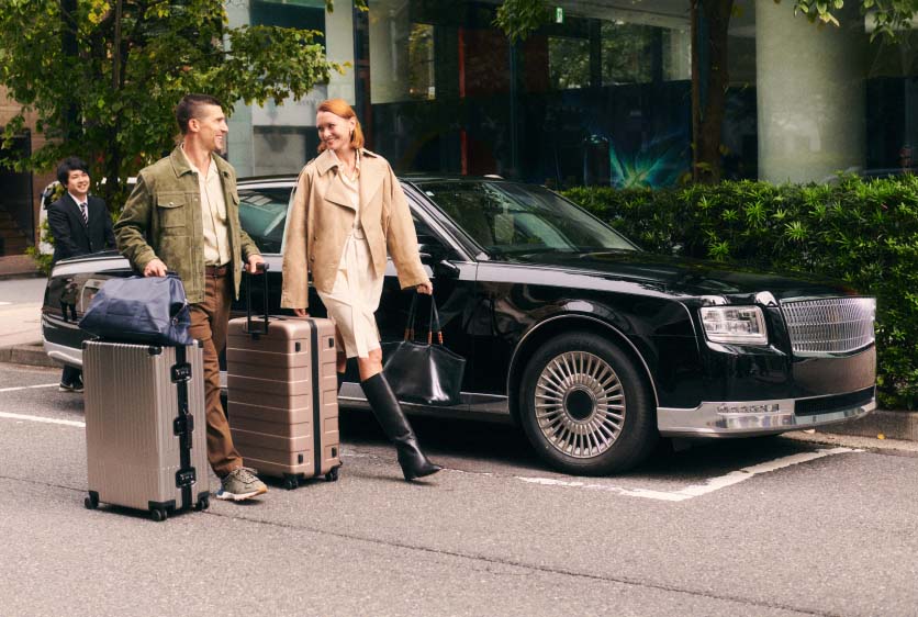 Two people walking on a street in Japan with luggage, in front of a black luxury car.