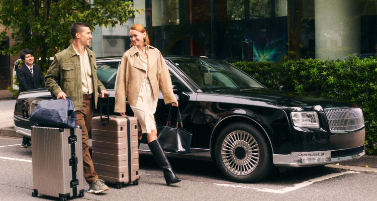 Two people walking on a street in Japan with luggage, in front of a black luxury car.