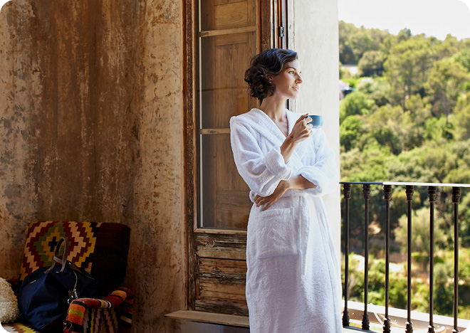 Woman in a robe standing on a balcony overlooking a green hillside.