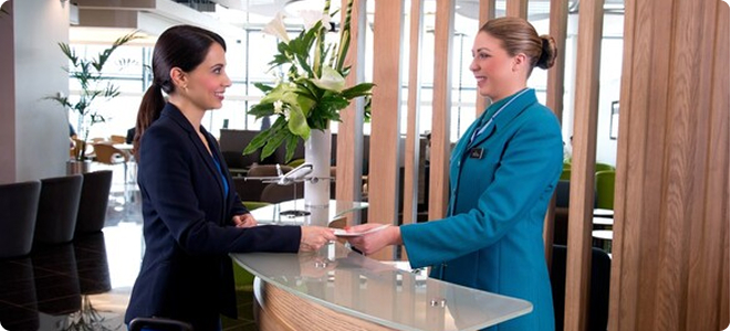 Traveler receives assistance at an airport lounge reception desk while learning about AerClub travel rewards benefits.