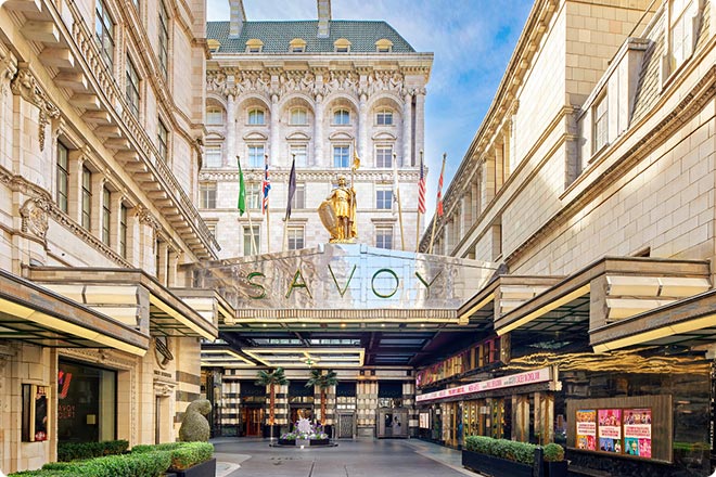 Exterior view of The Savoy Hotel entrance in London, showing the illuminated canopy and Savoy Theatre marquee along the street.