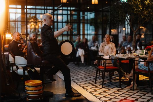 Performer playing drum and speaking to audience.