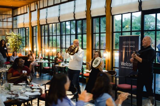 Performer playing drums and speaking to seated guests indoors.