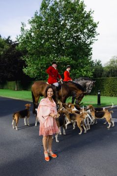 Riders on horseback with hounds on estate path.