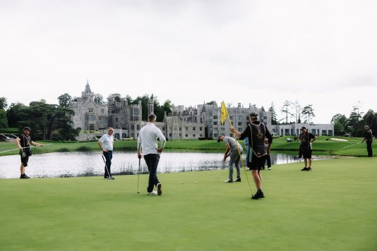 Golfers walking across a green near water.
