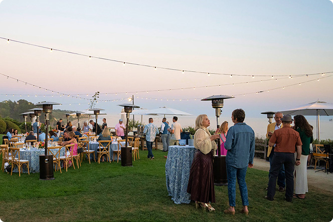 Guests gathered outdoors on a lawn for a reception with string lights and ocean views at sunset