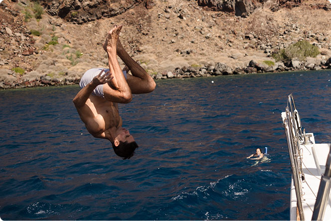Person diving off a boat into clear blue water near a rocky coastline