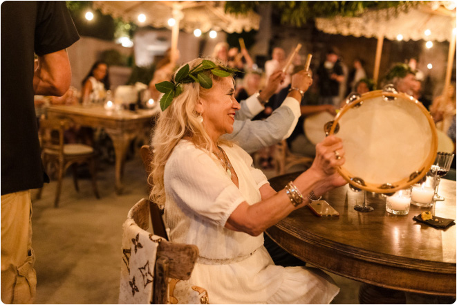 Guest seated at a table holding a tambourine during a lively indoor Greek cultural gathering
