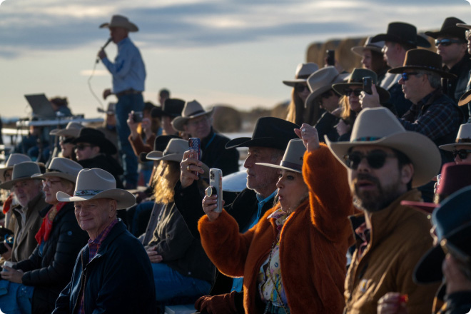 Crowd seated in outdoor bleachers watching a rodeo event, many wearing cowboy hats
