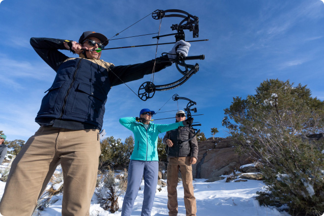 Person aiming a bow outdoors in a snowy landscape with two others standing nearby