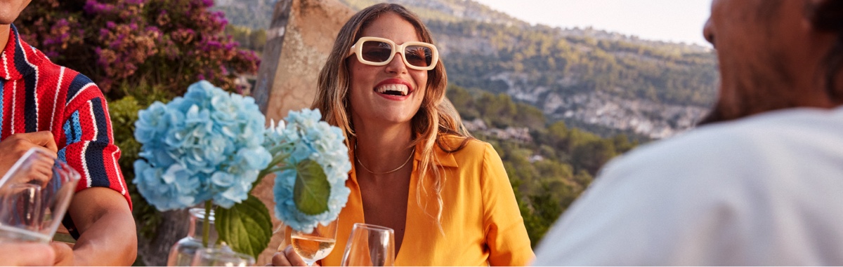 A stylish woman and her friends sit around an outdoor dining table, eating and drinking